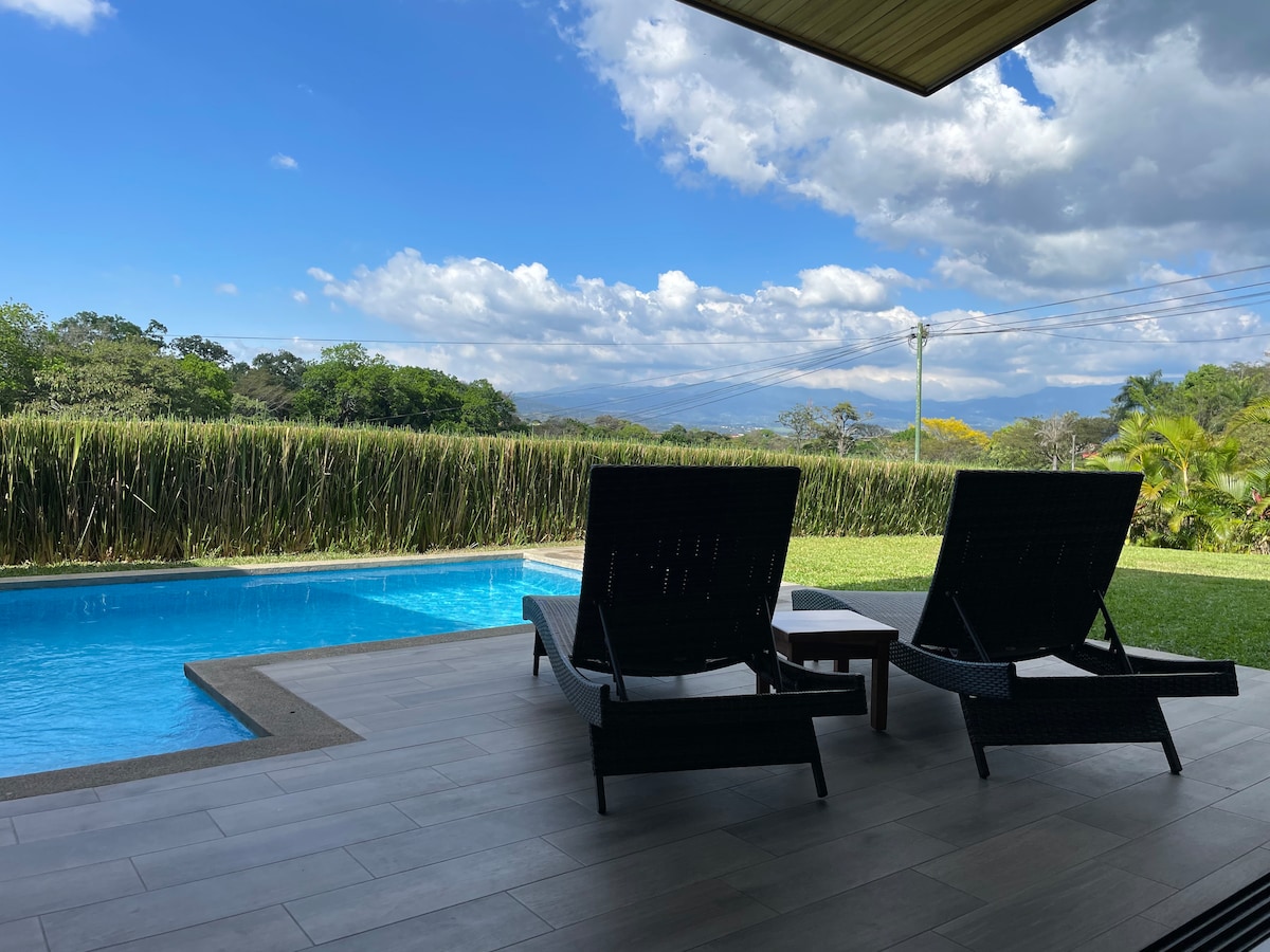 An outdoor area features two lounge chairs positioned alongside a serene swimming pool, with expansive valley views in the background. Lush greenery and distant mountains are visible under a partly cloudy sky, creating a peaceful atmosphere for relaxation.
