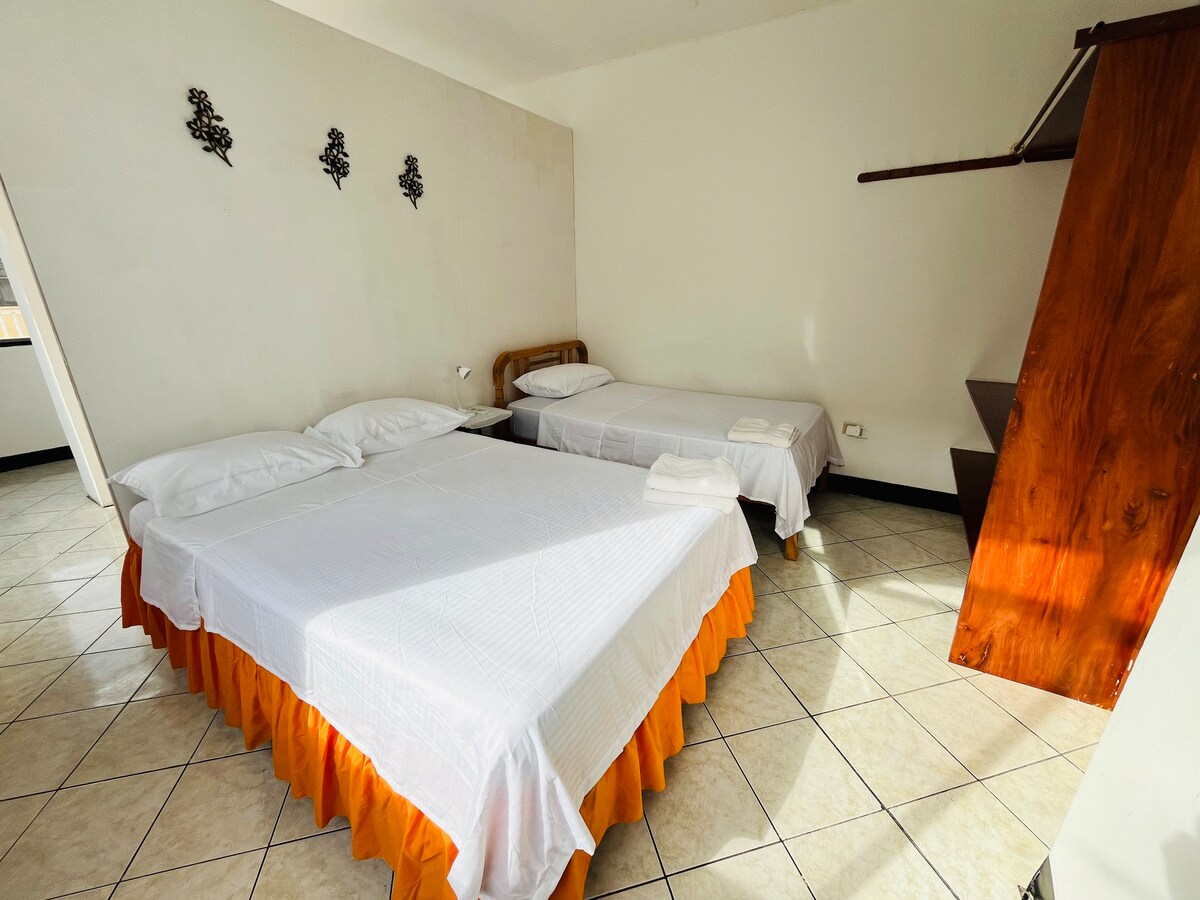 The sleeping area features two semi-independent beds, one larger and one smaller, both dressed in white linens with an orange bed skirt. A simple wooden shelf is mounted on the wall, and two decorative wall hangings add minimal design.