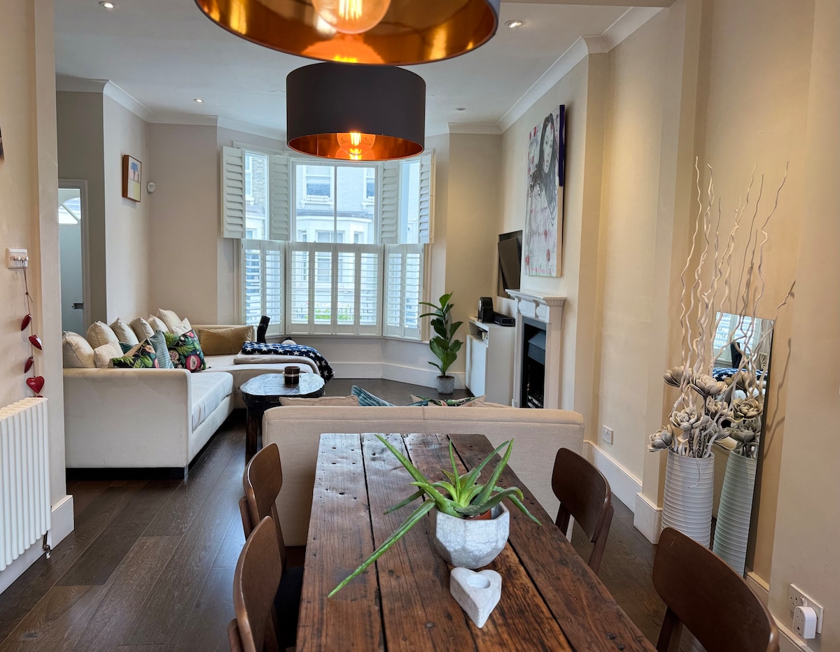 The open living and dining area features beautiful wooden floors and a large vintage wooden dining table with chairs. A cozy seating area includes two large sofas and a stylish coffee table, framed by natural light pouring in through expansive windows adorned with shutters.