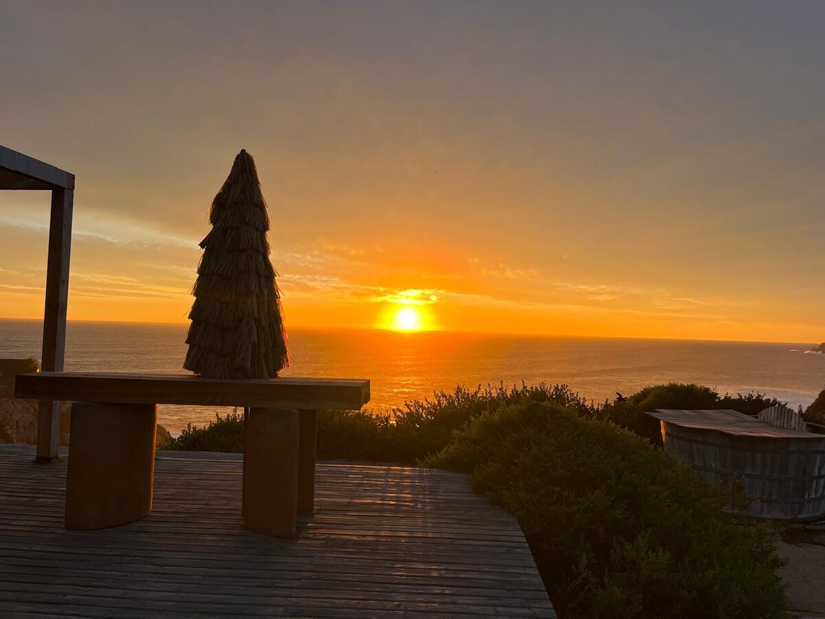 A silhouette of a structured tree-shaped feature stands prominently against the backdrop of a vibrant sunset over the ocean. The warm hues of orange and yellow reflect on the water, creating a serene atmosphere. The surrounding landscape consists of lush greenery and wooden decking.