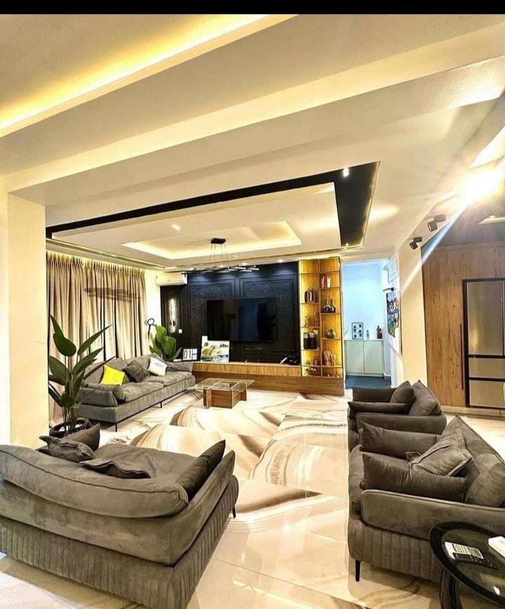 A modern living area features two plush sofas and a cozy armchair arranged around a coffee table. A large flat-screen TV is mounted on a stylish wall unit illuminated by soft lighting. Floor-to-ceiling windows with sheer curtains allow natural light to fill the space.