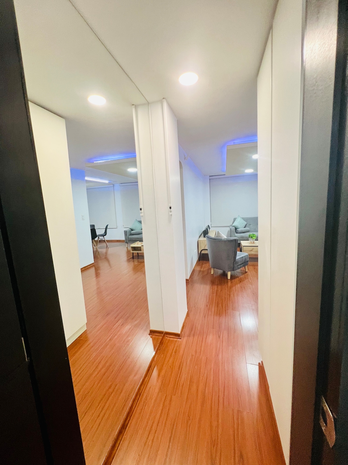 A view of a spacious hallway leading to a bright living area. The polished wooden floor reflects the warm light from the ceiling fixtures. Soft seating is visible in the background, complemented by contemporary decor and large windows dressed with light curtains.