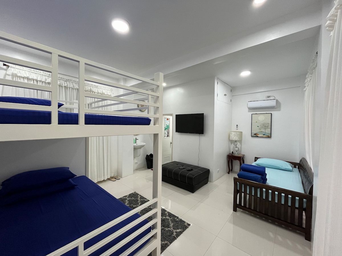 A spacious room features two sleeping areas, including a lofted bunk bed with blue bedding and a lower bed with additional light blue linens. A dark ottoman and a wall-mounted television create a comfortable space, complemented by soft lighting and white curtains.