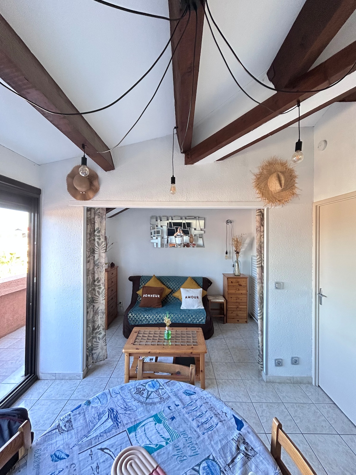 A bright living space is featured, showcasing exposed wooden beams overhead. A cozy bed with decorative pillows is visible in the background, while a wooden dining table occupies the foreground. Natural light floods the room through the glass doors leading to a balcony.