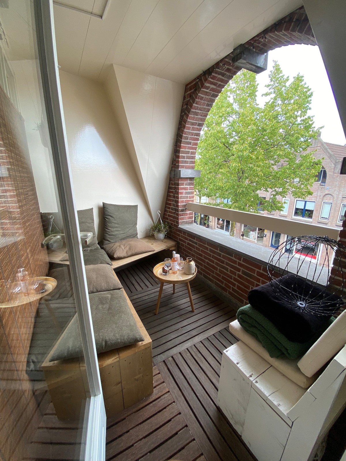 An outdoor balcony features a wooden deck and a combination of cushioned seating along the wall. A small circular table rests at the center, accompanied by a selection of amenities. Green foliage can be seen just outside, providing a natural view.