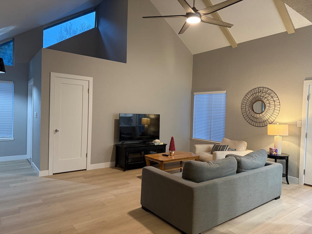 A spacious living area features a comfortable light gray sofa and a wooden coffee table. A flat-screen TV sits on a stand, with a lamp illuminating the corner. Natural light enters through windows, while a decorative mirror enhances the wall's simplicity.