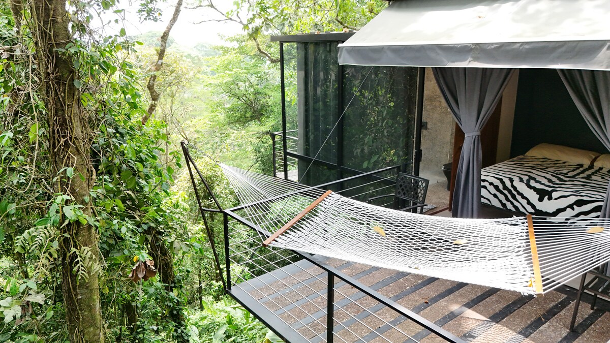 A hammock is suspended on a deck, surrounded by lush greenery. The structure features an entrance with floor-to-ceiling curtains leading to a cozy sleeping space with a zebra-patterned bed. The serene environment offers a sense of tranquility within the trees.