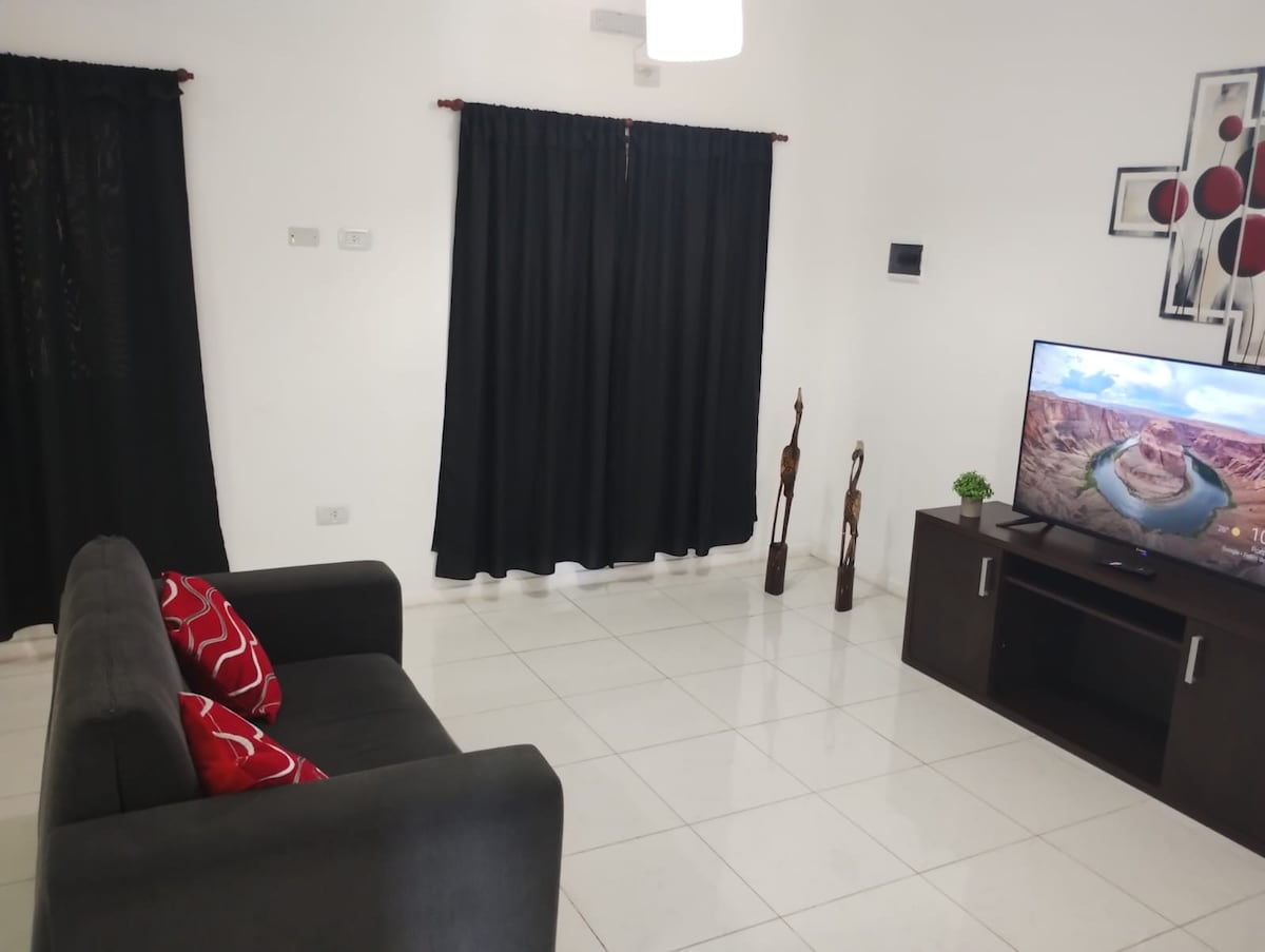 A living area features a dark sofa adorned with red pillows, positioned near a television set on a dark furniture unit. Two tall decorative sculptures stand alongside the wall. Black curtains cover the windows, contrasting with the bright tiled floor.