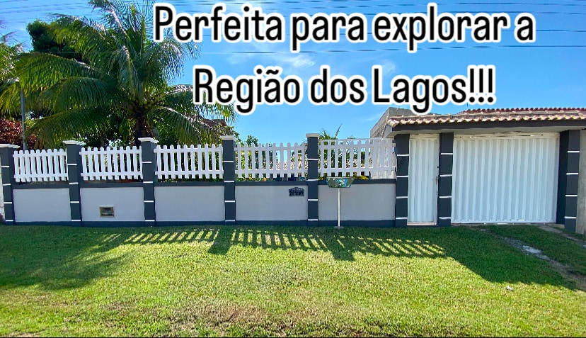 A well-maintained exterior of a property is showcased, featuring a white picket fence and a manicured lawn. The house is visible behind the fence, with palm trees adding to the pleasant surroundings. Bright sunlight enhances the welcoming appearance of the entrance.