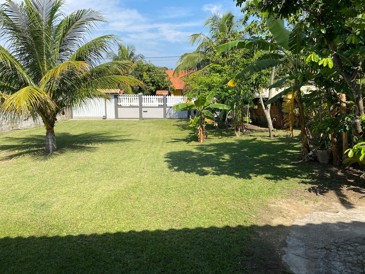 A spacious backyard is surrounded by lush greenery, featuring several palm and fruit trees. The green grass is well-maintained, providing an open area for outdoor activities. A white fence partially encloses the space, ensuring privacy. Clear skies and sunlight create a welcoming ambiance.