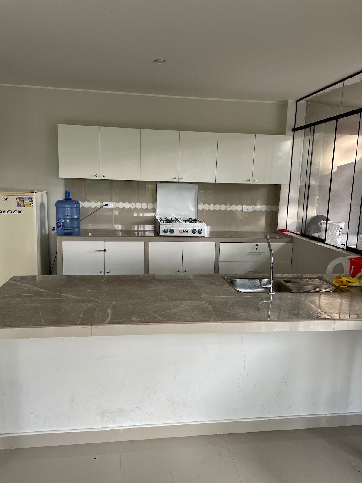 A modern kitchen features white cabinetry and a spacious countertop made of light-colored stone. An upright refrigerator, water dispenser, and a gas stove with an overhead vent are visible. Natural light streams in through large windows, enhancing the functional layout.