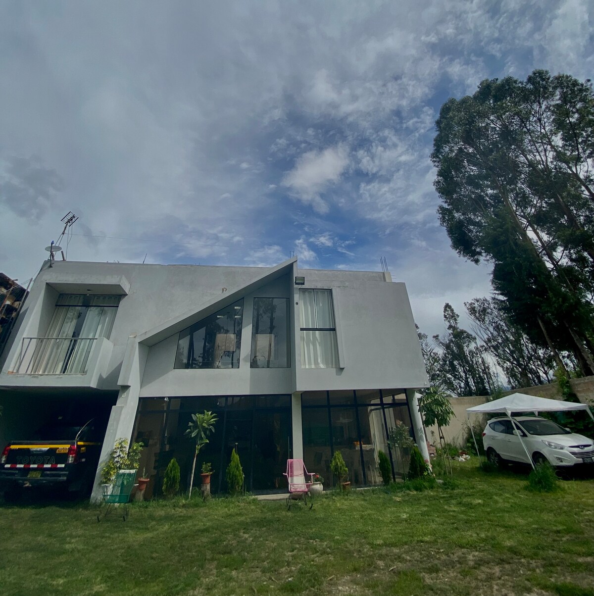 The exterior of a spacious house is presented, featuring large windows that reflect natural light. Surrounding greenery includes trees and a well-maintained lawn. A driveway accommodates vehicles, while a covered area offers additional parking. Cloudy skies provide a calm backdrop to the structure.