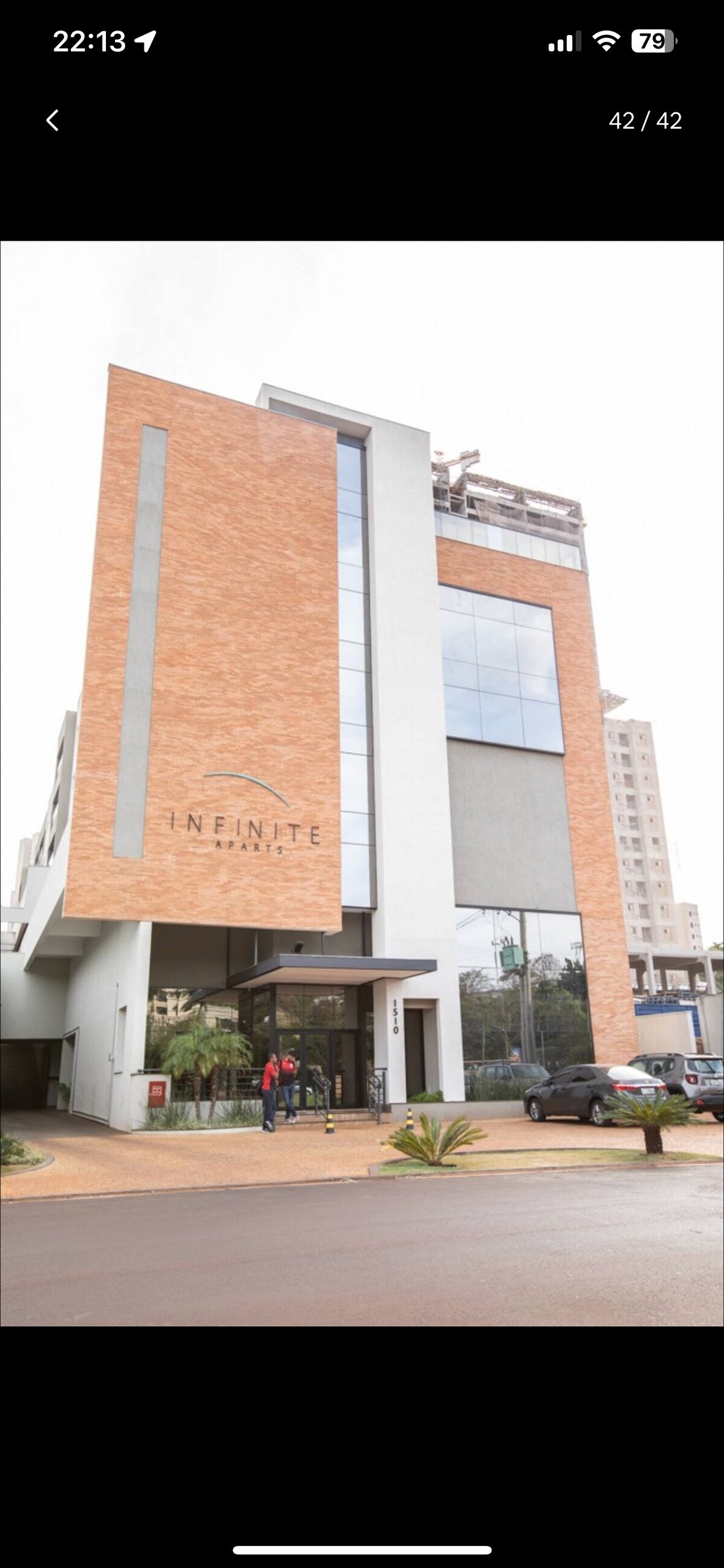 The image showcases a modern building with a distinctive brick facade and large glass windows. The entrance features a welcoming canopy, and potted plants are arranged along the pathway, enhancing the accessibility of the main entrance.