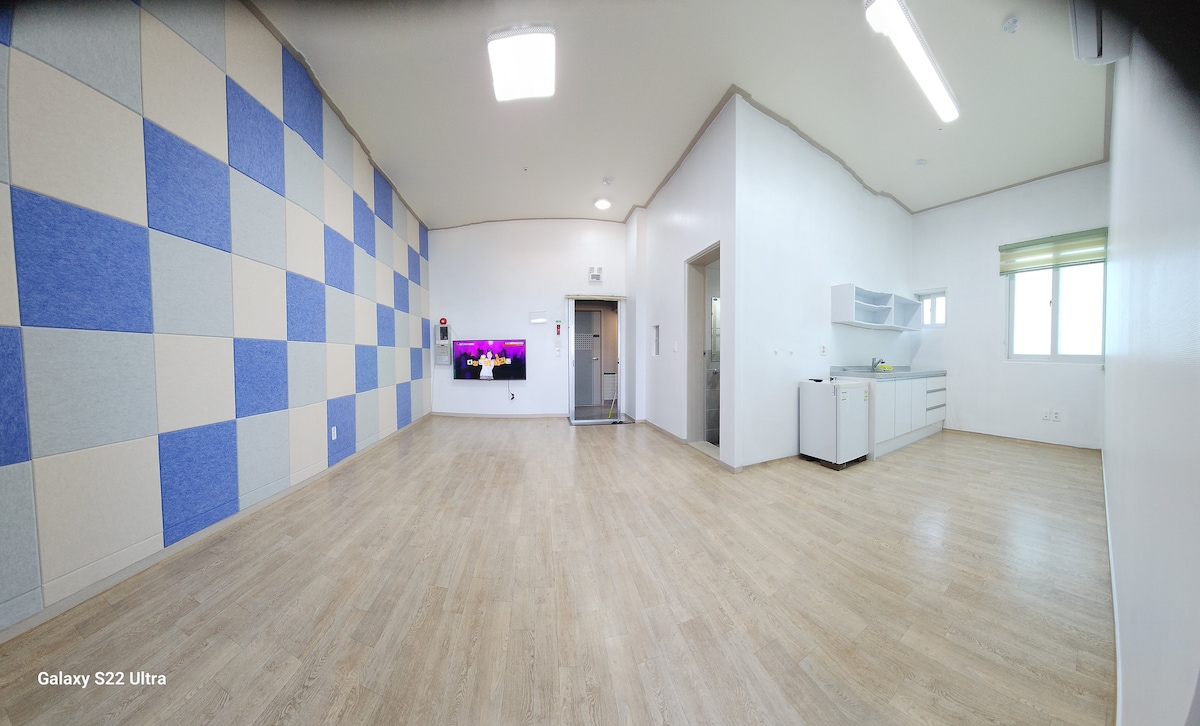 A spacious room is presented with patterned blue and gray walls. Light wood flooring complements a modern kitchenette on one side. A large window allows natural light to fill the area, while a flat-screen TV is mounted on the wall opposite the kitchenette.