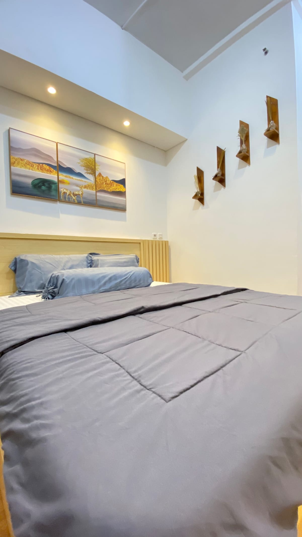 A cozy sleeping area features a spacious bed with light grey bedding, complemented by decorative pillows. Wall-mounted art pieces with scenic landscapes enhance the room's ambiance. Natural light enters through the room's upper section, creating a bright and airy feel.