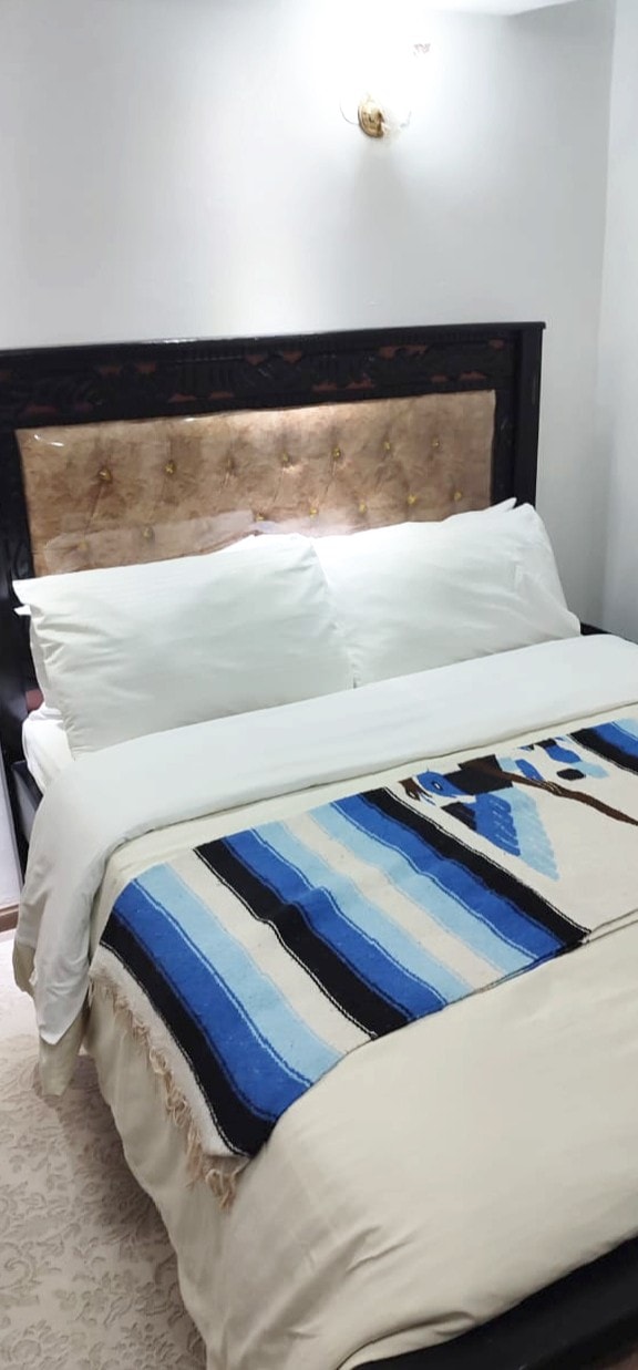 A cozy sleeping area features a spacious bed with plush white pillows and a decorative blanket showcasing blue and beige stripes. The headboard is upholstered in a neutral tone, adding a touch of elegance to the room's simple design.