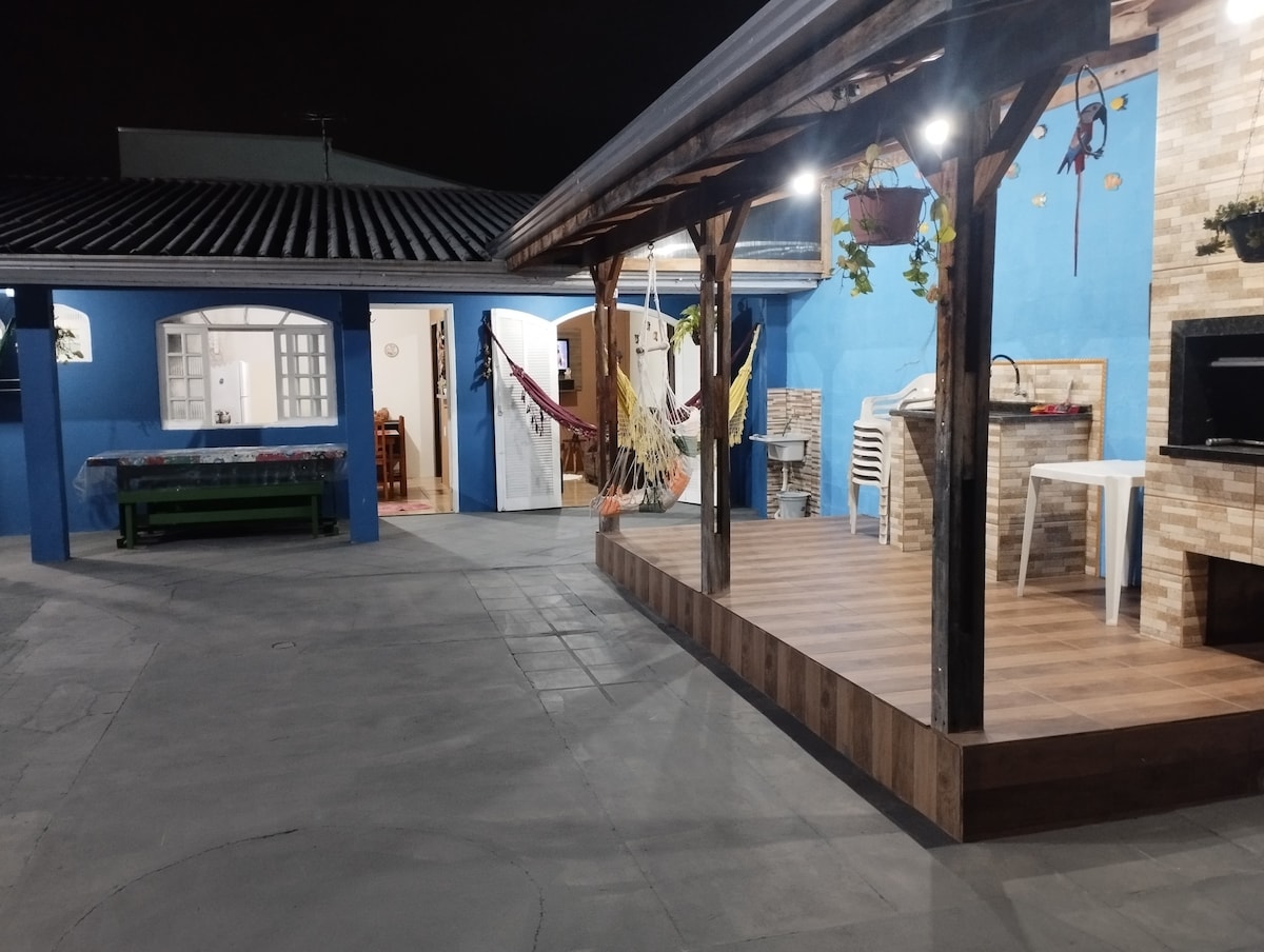 An outdoor area features a wooden deck with a grill and seating. Hammocks are suspended for relaxation. A blue wall and a stone fireplace add character to the space. The entrance leads to the kitchen and dining area, enhancing the inviting, communal atmosphere.