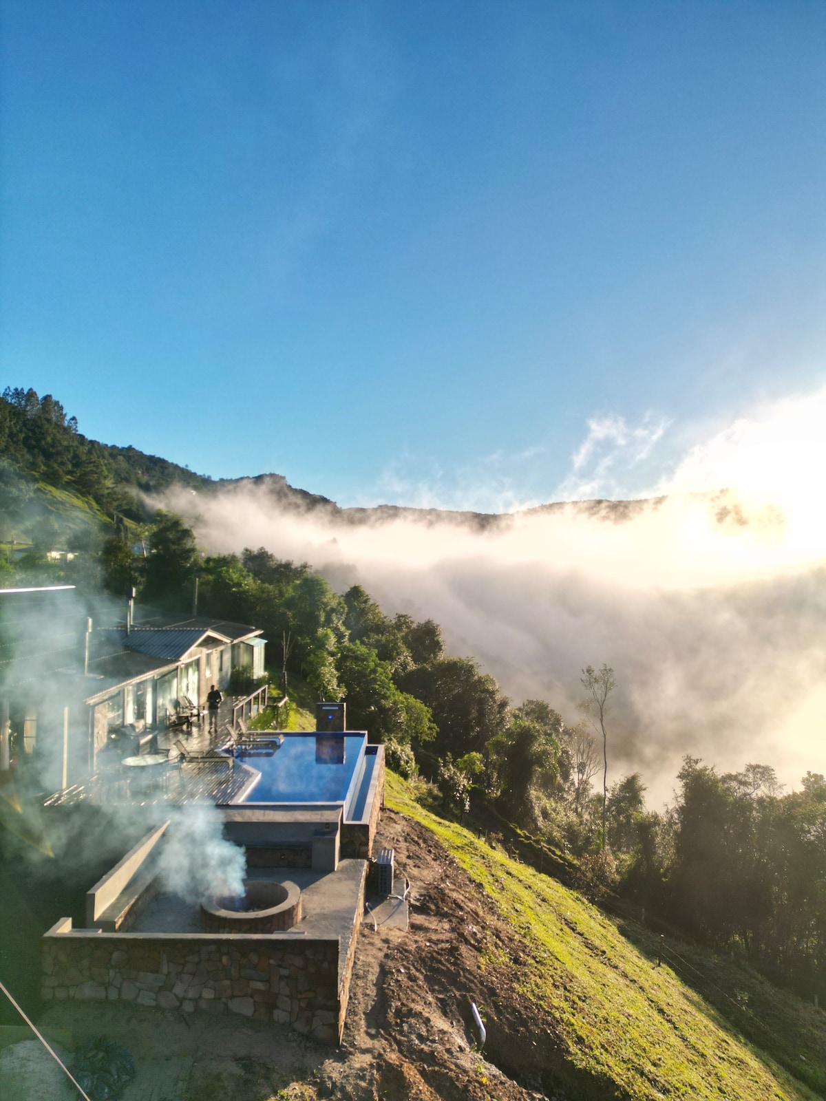 The image showcases a stunning landscape featuring a swimming pool perched on a hillside, surrounded by lush greenery. Mist rolls through the valley below, while the sun casts a warm glow over the scene. The structure of the property appears modern, integrating seamlessly with its natural surroundings.