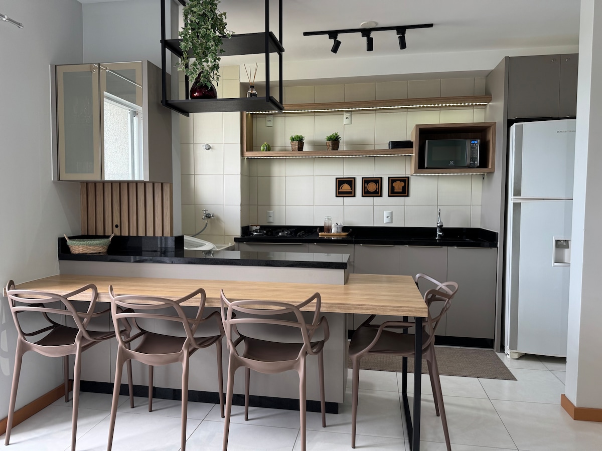 The kitchen features modern cabinetry, a spacious counter, and essential appliances, including a microwave and refrigerator. A dining table with six chairs is positioned centrally, complemented by decorative elements on shelves above. Natural light enters through a nearby window, enhancing the open layout.