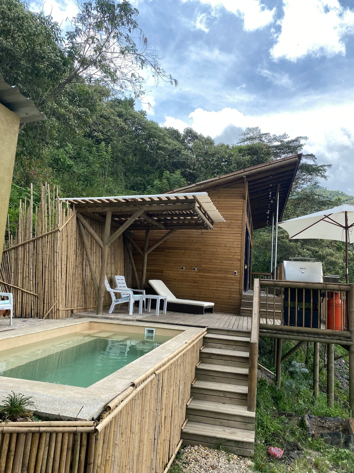 A wooden cabin is set against a lush mountainous backdrop. A small private pool is bordered by bamboo fencing, with two white lounge chairs positioned nearby. Steps lead from the pool area to a raised deck, featuring a large umbrella for shade.