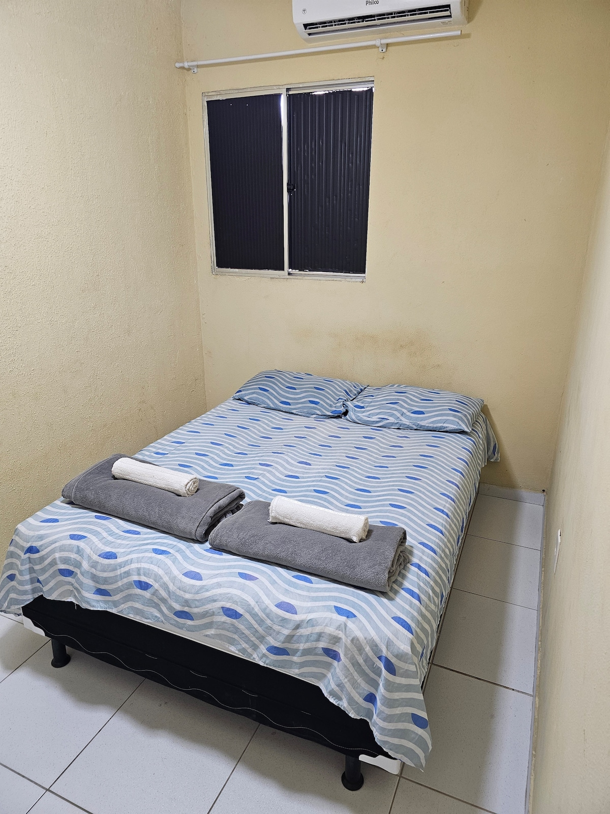 A cozy bedroom is featured, featuring a spacious bed dressed in a patterned blanket. Two towels are neatly arranged on the bed. A single window provides natural light, while an air conditioning unit is installed above, ensuring comfort.