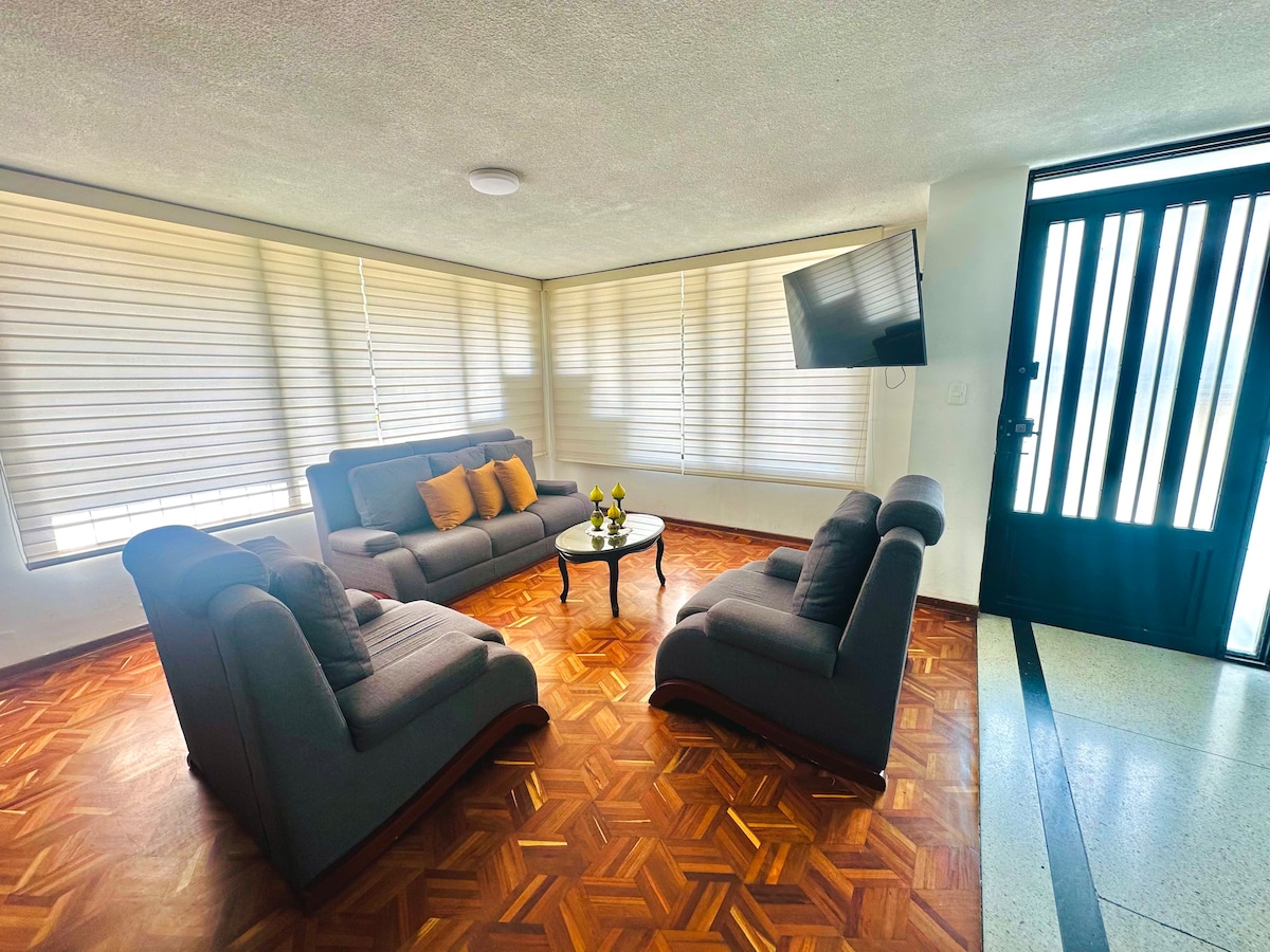 A spacious living area is filled with natural light from large windows covered with horizontal blinds. Comfortable seating is provided by a gray sofa with yellow accent pillows, complemented by an elegant coffee table placed in the center. A flat-screen TV is mounted on the wall.