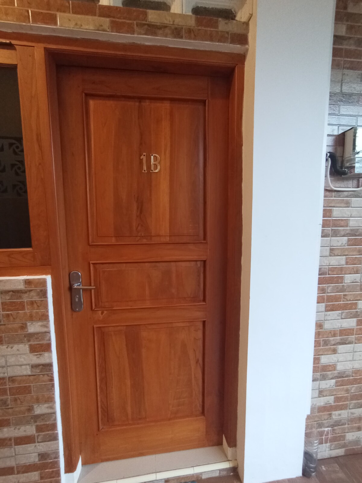 A wooden door marked with the number '1B' is shown, featuring a classic design with clean lines and a polished finish. The door is framed by a textured wall with a combination of light and dark tiles, reflecting a warm and inviting aesthetic.
