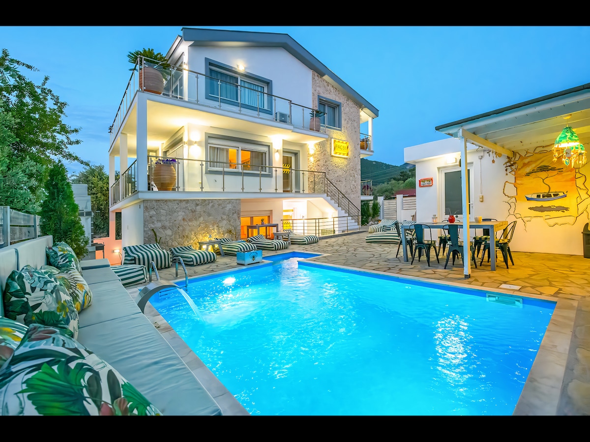 An inviting outdoor space showcases a well-lit pool surrounded by comfortable seating. The structure features multiple levels with a blend of stone and modern design. A dining area with colorful decor is visible nearby, enhancing the atmosphere for family gatherings.
