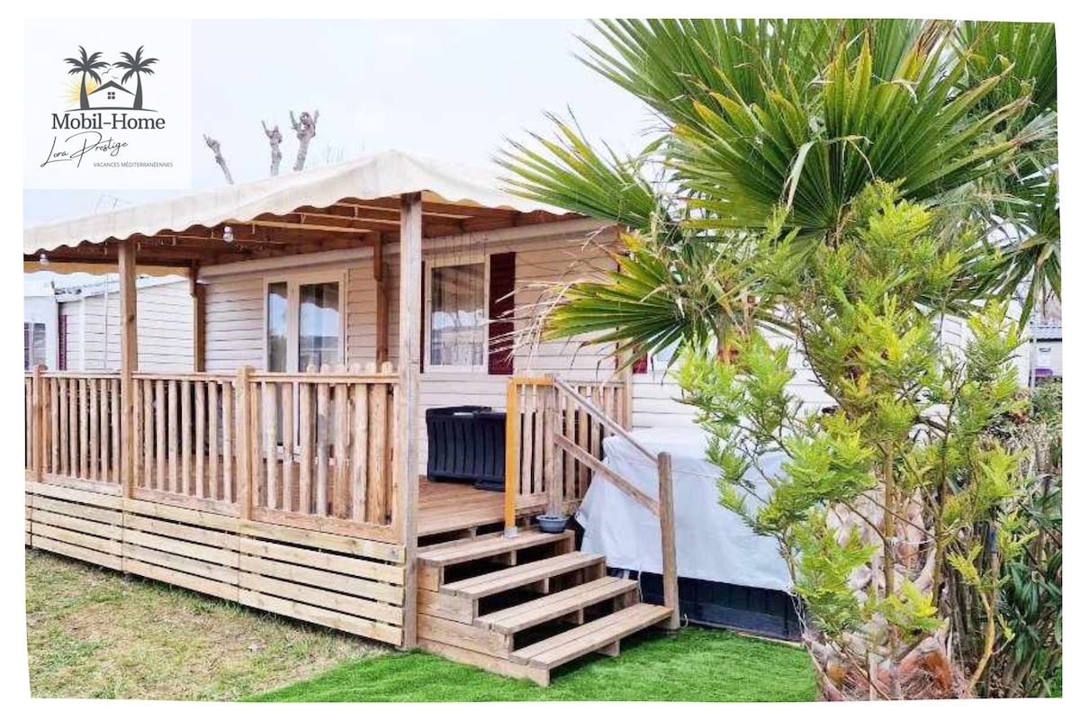 A mobil-home features a wooden deck with steps leading up to the entrance. Lush greenery surrounds the exterior, providing a natural setting. A covered terrace offers shade, while the building's exterior is finished with a light color, contributing to a spacious appearance.