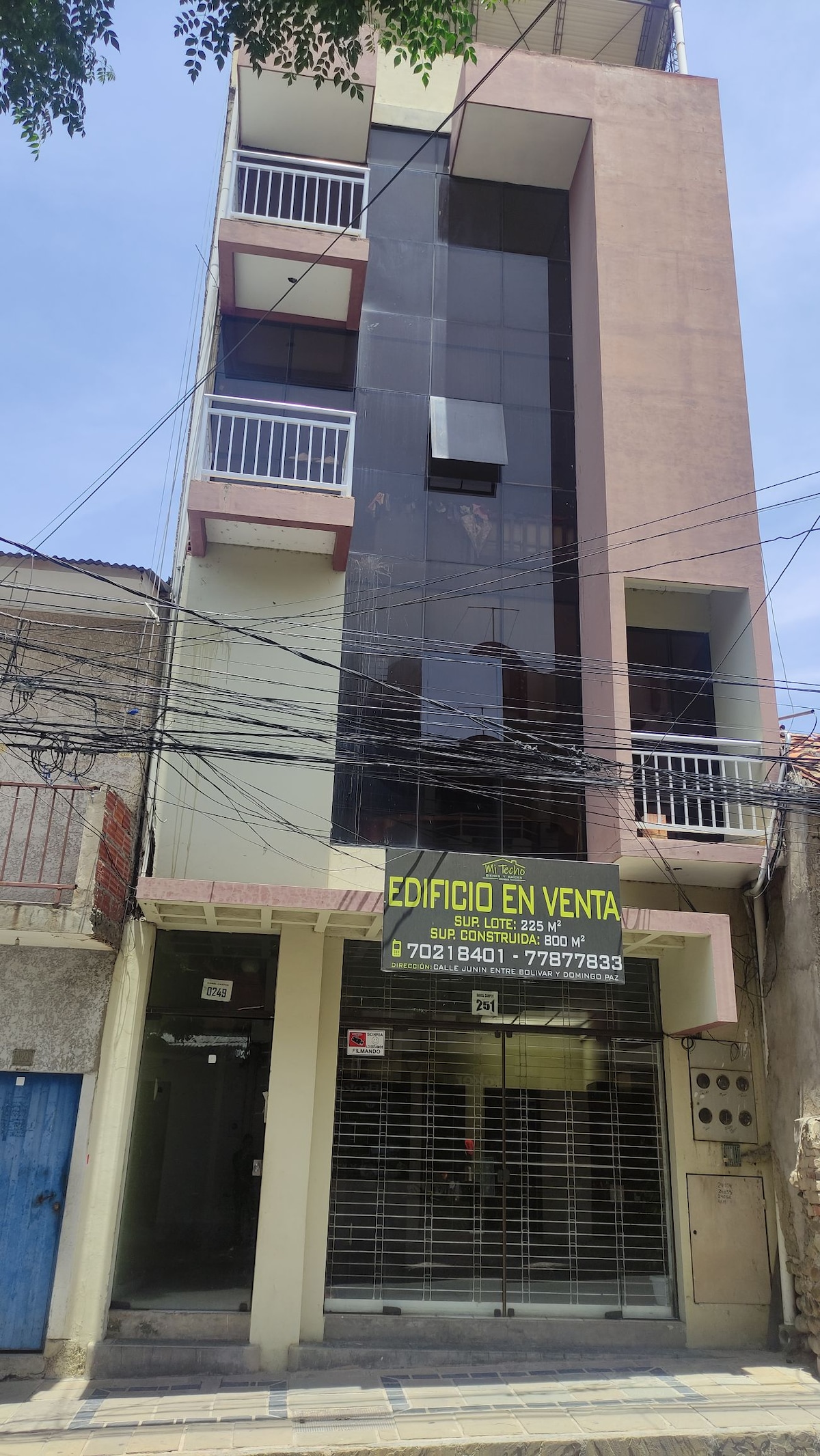 The exterior of a modern multi-story building is visible, featuring a blend of glass and concrete elements. A prominent sign indicates property for sale. The entrance is secured with glass doors, and various electrical wires are seen overhead, framing the structure.