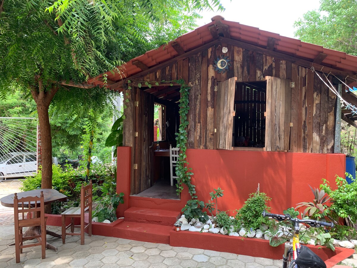 A charming wooden structure with a red-painted base is surrounded by lush greenery. A small table and chairs are positioned on the outdoor patio, creating a cozy seating area. Vines climb the exterior walls, adding natural elements to the inviting facade.