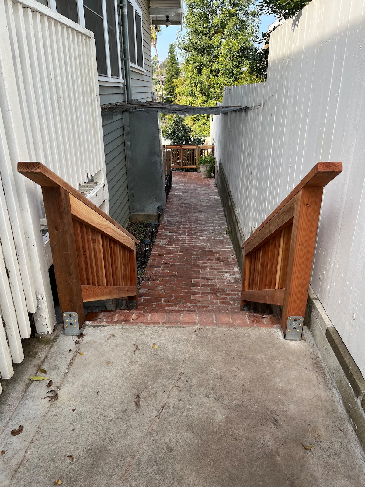 A brick pathway winds between two wood fences, leading towards a peaceful garden area. A set of wooden stairs is visible at the beginning of the path, guiding the way with sturdy handrails on either side.