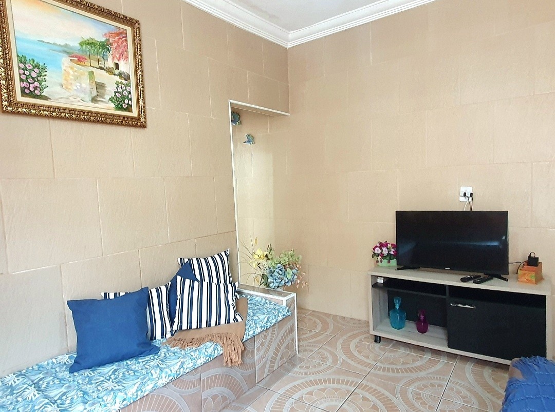 A living area is presented with light-colored tiled flooring. A television is positioned on a minimalistic cabinet, while a mirror reflects the space. Comfortable seating is offered with a patterned sofa and cushions, enhancing the overall inviting ambiance.