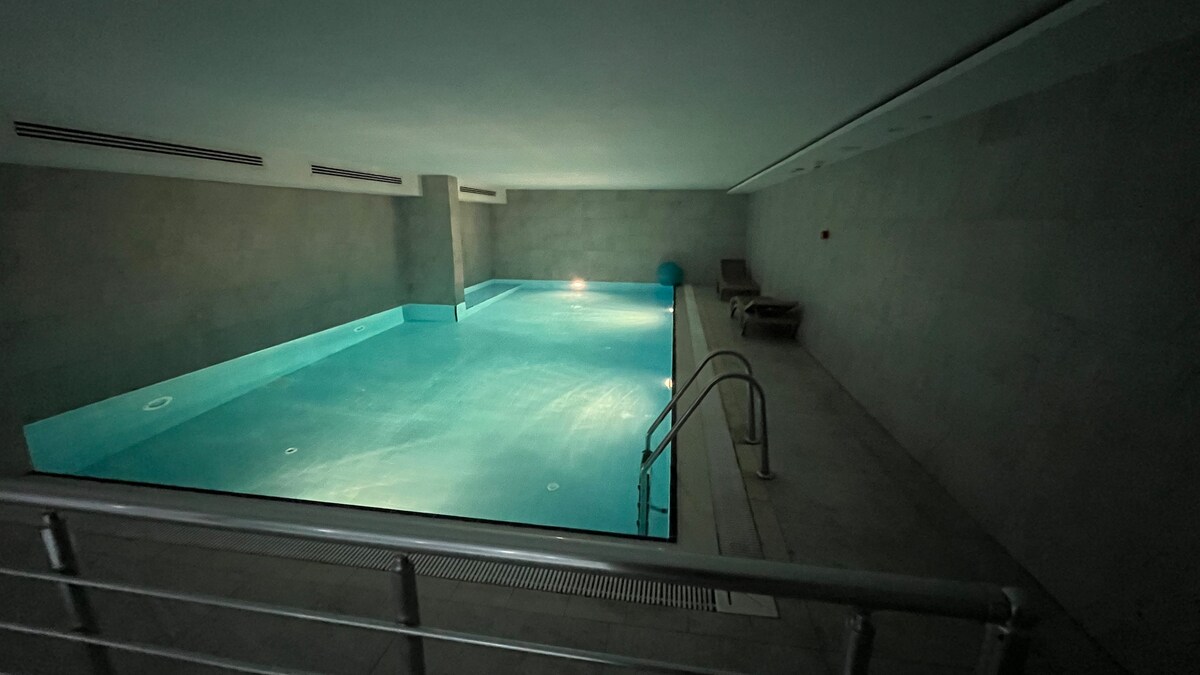 An indoor swimming pool is illuminated with soft lighting, creating a serene environment. The clear water reflects the light, and metal handrails lead into the pool area. Lounge chairs are positioned nearby, emphasizing a tranquil space for relaxation.