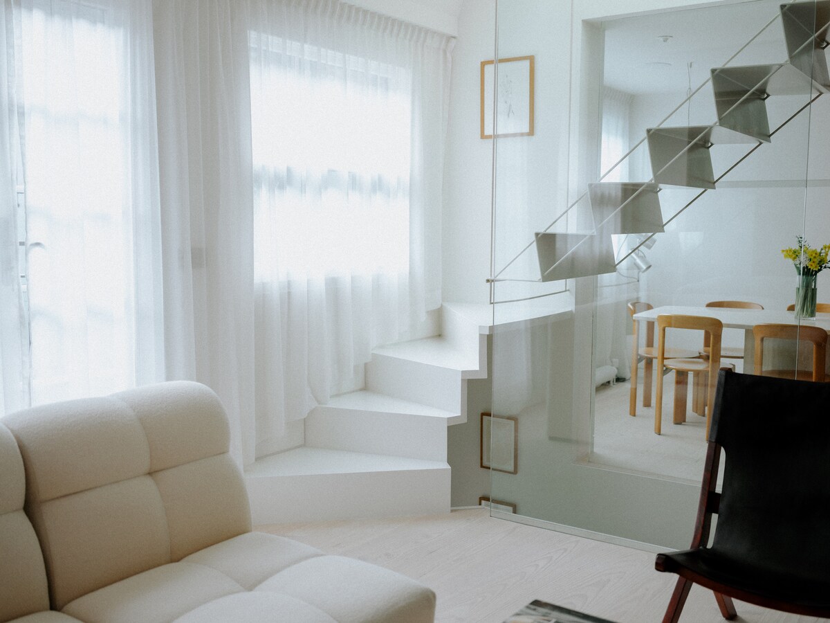 A bright living area is highlighted by large windows adorned with sheer white curtains, allowing natural light to fill the space. A comfortable sofa is positioned nearby, with a staircase leading to an upper level. A dining area with a wooden table and chairs is visible through a glass partition.