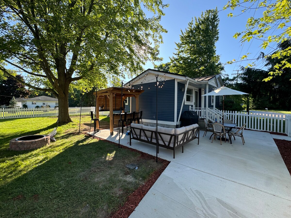 The outdoor space features a patio area with a wood firepit surrounded by chairs. A shaded gazebo is visible alongside a spacious seating arrangement. Lush green grass and a white picket fence enhance the serene environment, complemented by shaded trees providing a cooling effect.