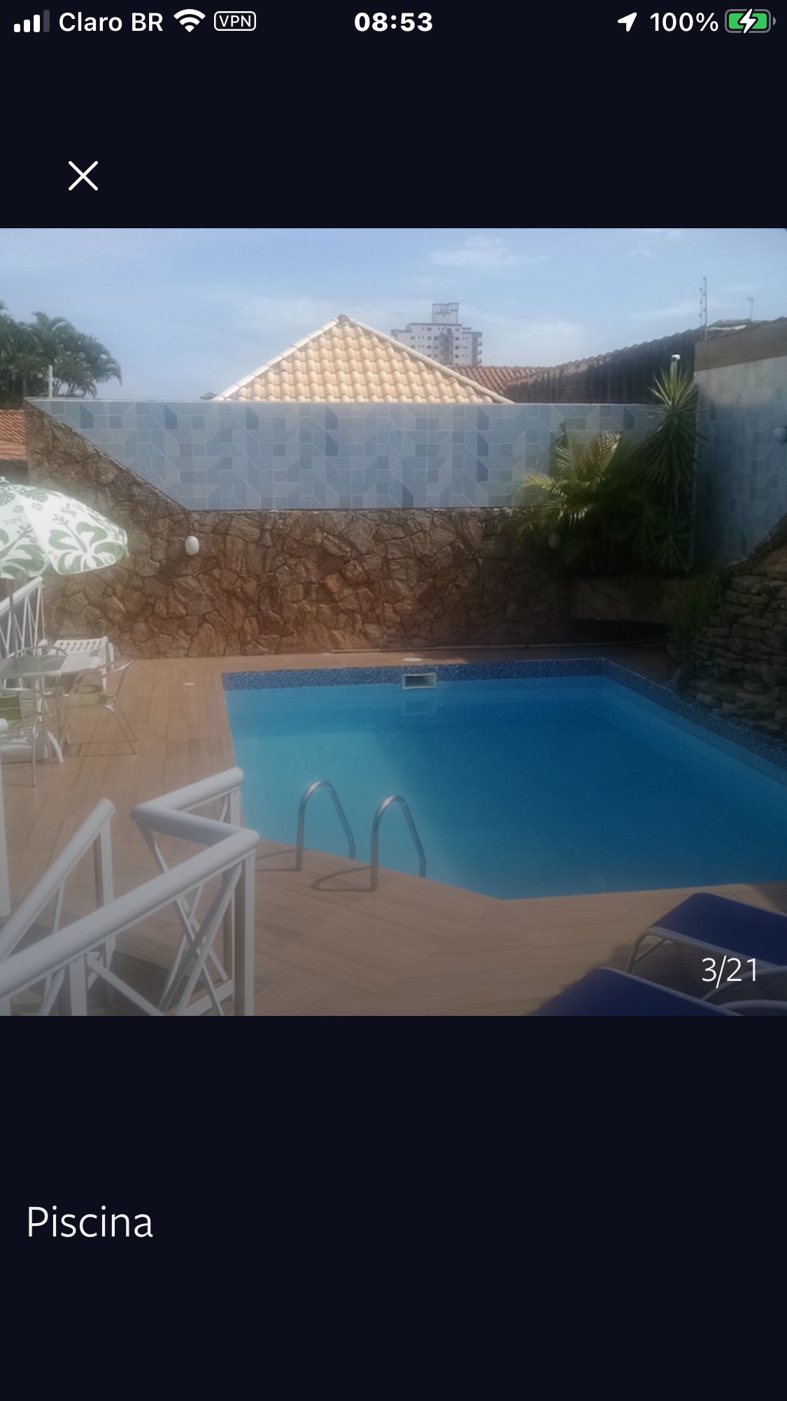 A rectangular pool with clear blue water is framed by a wooden deck. Two lounge chairs are positioned on one side, while decorative green and white umbrellas provide shade. The surrounding area features a stone wall and greenery, enhancing the outdoor space.