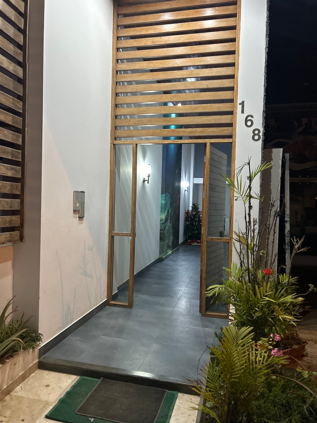 The entrance features a prominent wooden slatted design, allowing natural light into a hallway. The floor is tiled in dark gray, and potted greenery adds a touch of nature along the sides. The address number is displayed prominently beside the door.