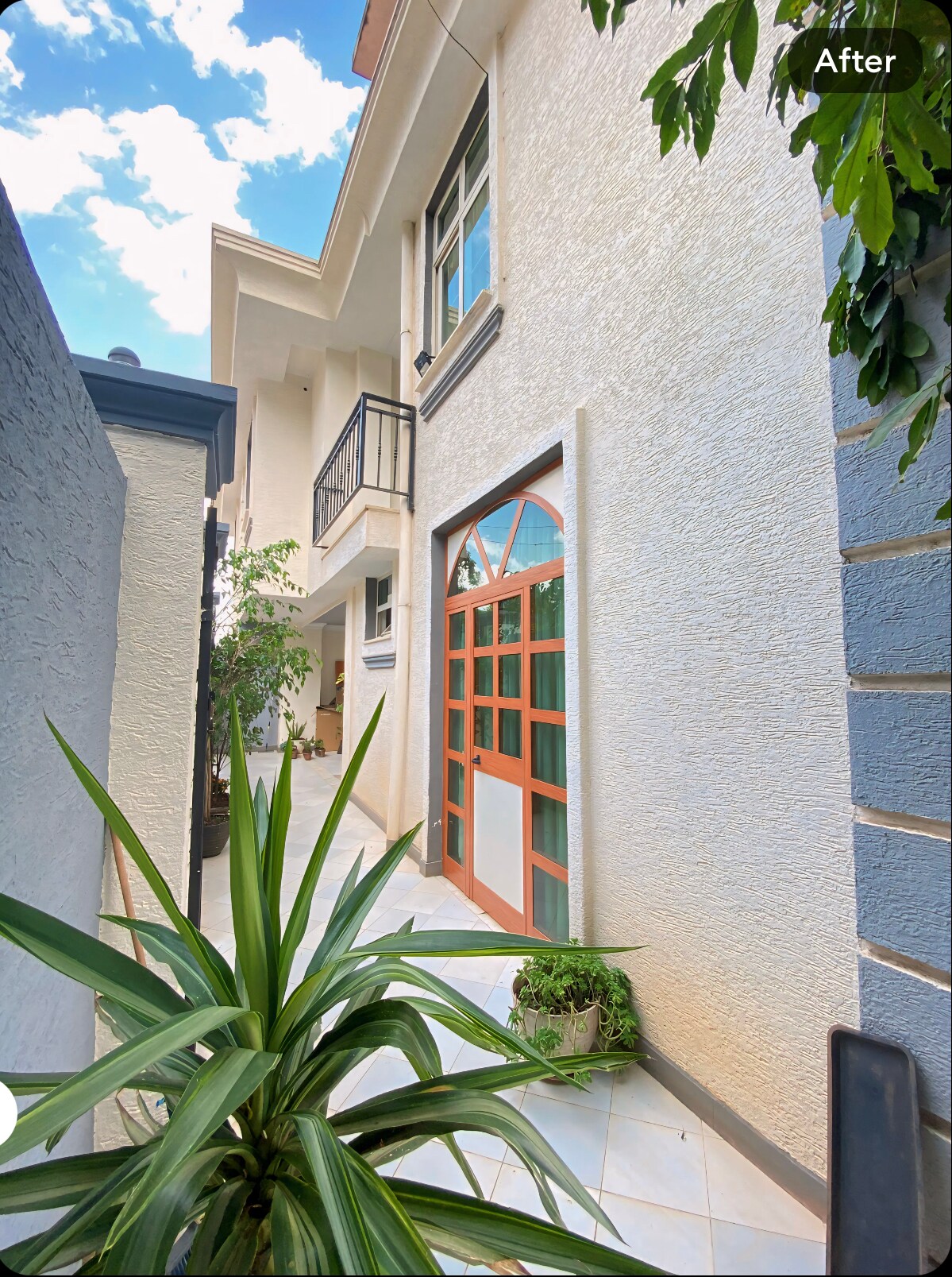 A pathway is framed by vibrant greenery leading to a well-lit entrance. The white exterior walls are complemented by a large arched window featuring wooden accents. Clear blue skies and scattered clouds can be seen above, enhancing the welcoming atmosphere.