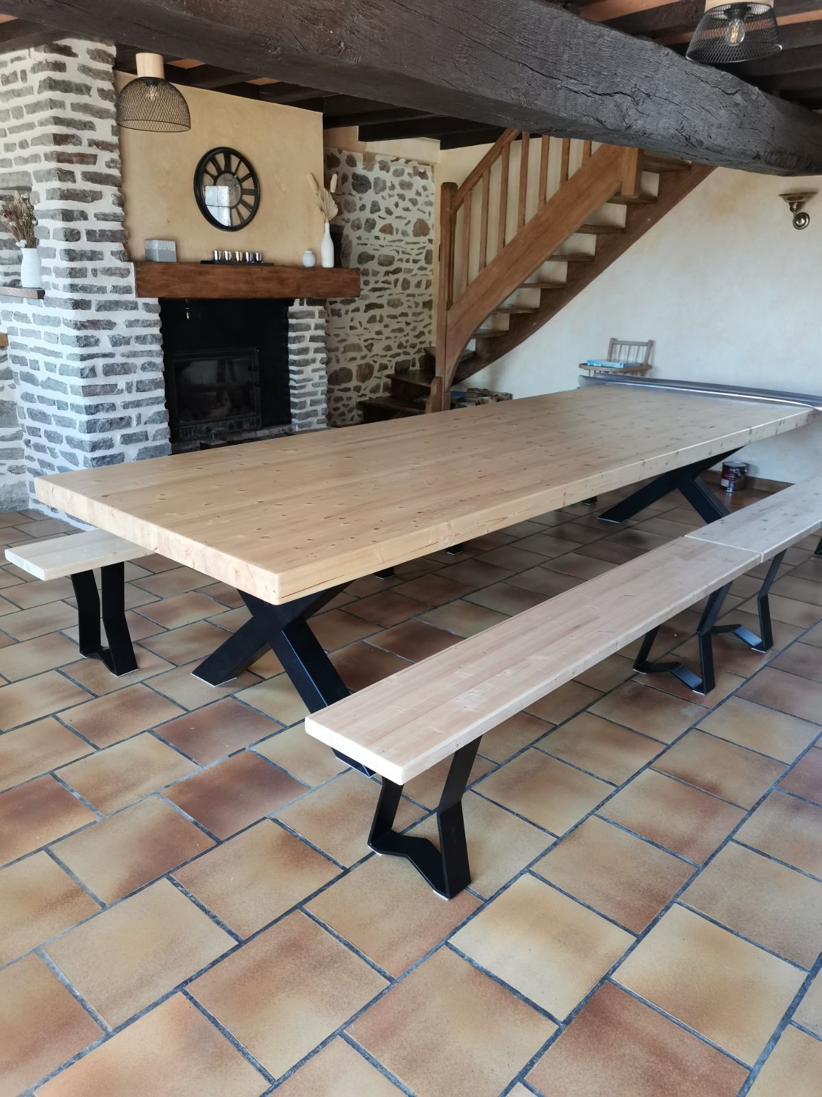 A wooden dining table with a natural finish is accompanied by matching benches, positioned in a room featuring rustic stone walls and a decorative clock. A staircase made of wood leads to an upper level, while a fireplace adds warmth to the space.