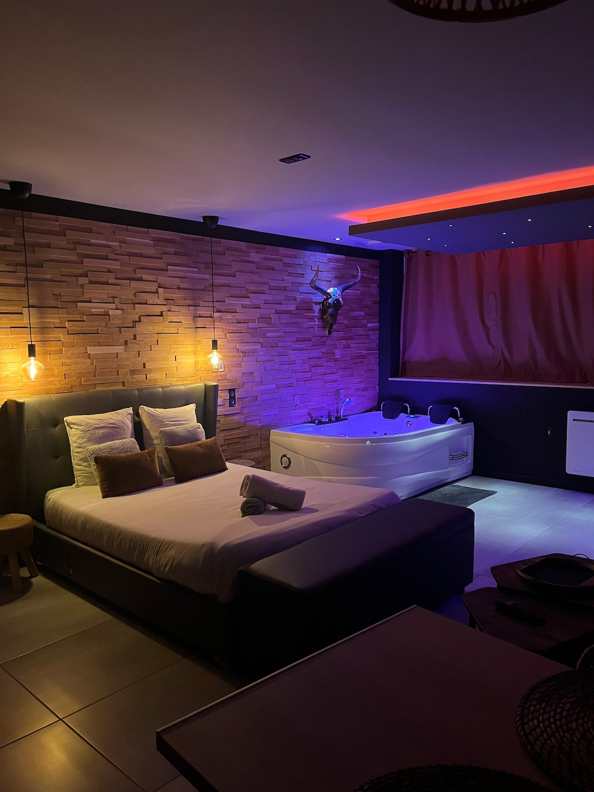 A comfortable sleeping area features a spacious bed with soft pillows. Warm pendant lights illuminate the textured brick wall, while a modern whirlpool tub sits nearby. Subtle accent lighting creates a relaxed ambiance, complemented by a cozy stool and installed air conditioning unit.