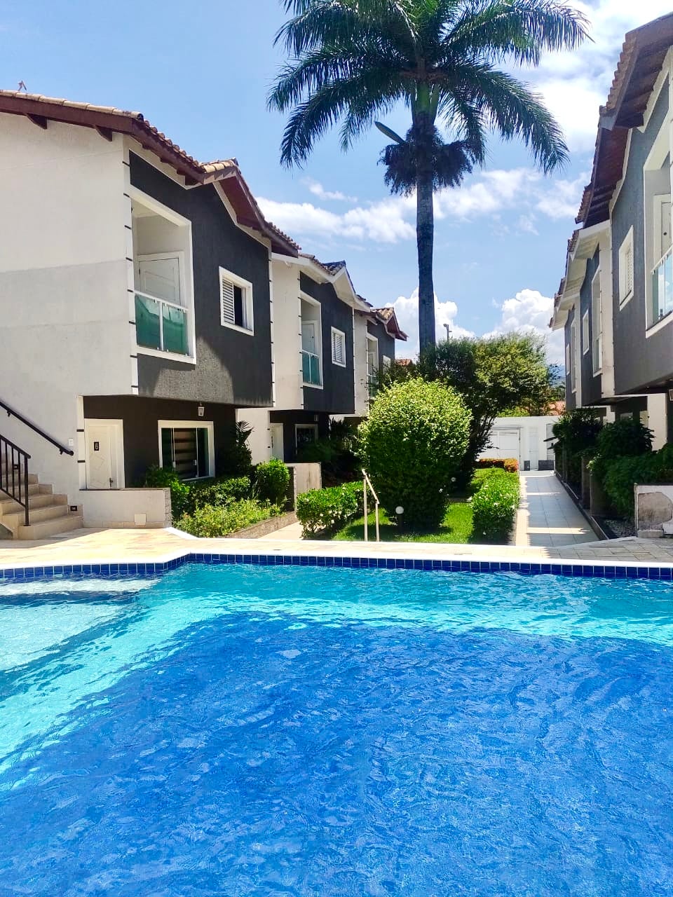 A clear view of the pool is presented, surrounded by well-maintained greenery and villas. The sun shines brightly, reflecting off the calm water. Lush palm trees are visible, complementing the tranquil atmosphere of the condominium complex.