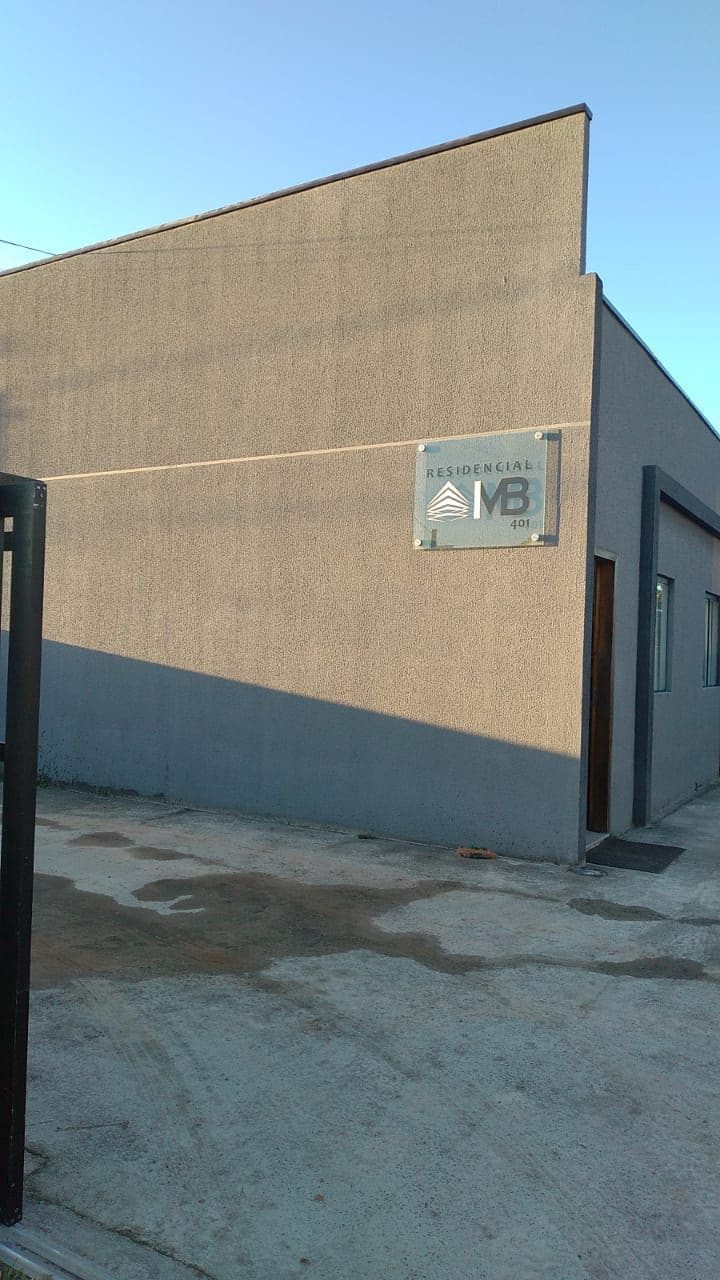 The exterior of a modern building is shown, featuring a gray facade. A clearly visible sign reads 'Residencial VB.' Natural light casts a shadow on the ground, highlighting the clean lines and simplicity of the structure.