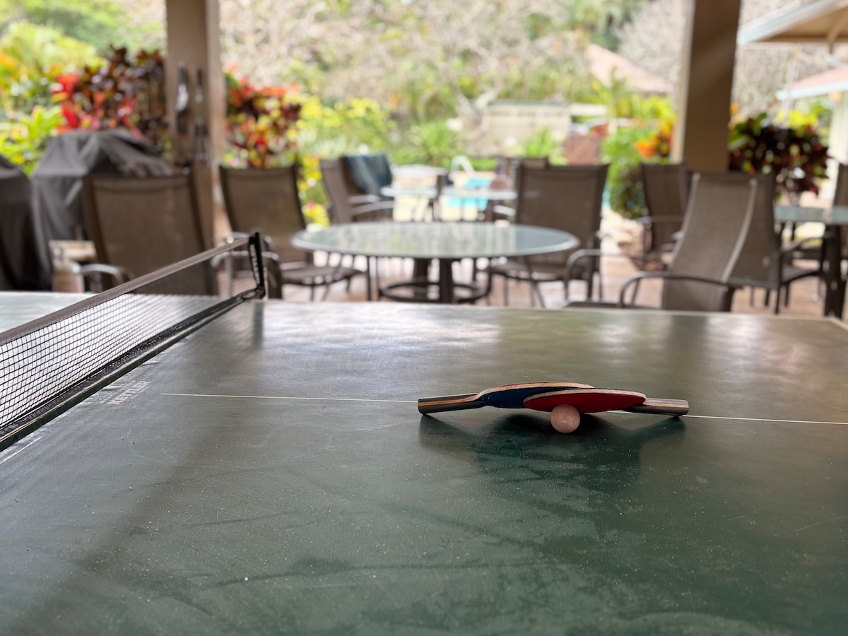 A ping pong table is situated in an outdoor area surrounded by tropical foliage and vibrant plants. A paddle and ball rest on the green table surface, while a gathering space with various chairs and tables is visible in the background.