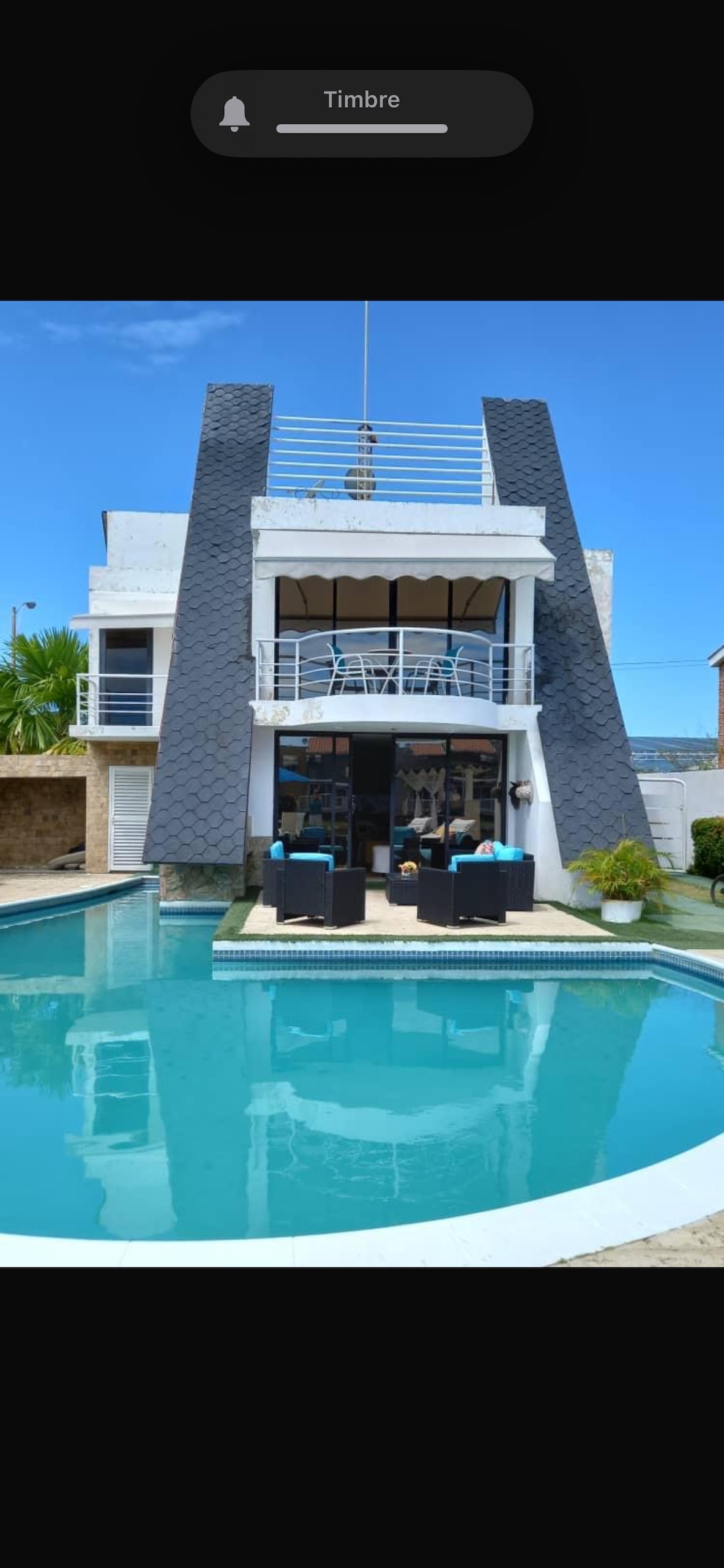 The contemporary villa features a sleek exterior with distinctive triangular pillars. A private swimming pool reflects the clear blue sky, while comfortable seating arrangements are visible on the terrace. Lush greenery surrounds the space, enhancing the overall ambiance of relaxation.