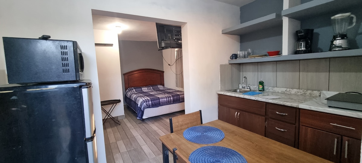 A clean and functional kitchenette is shown, featuring a refrigerator, wooden cabinets, and an assortment of small appliances including a coffee maker and blender. A dining table with two chairs is visible. Beyond, a cozy sleeping area with a patterned blanket on the bed can be seen.