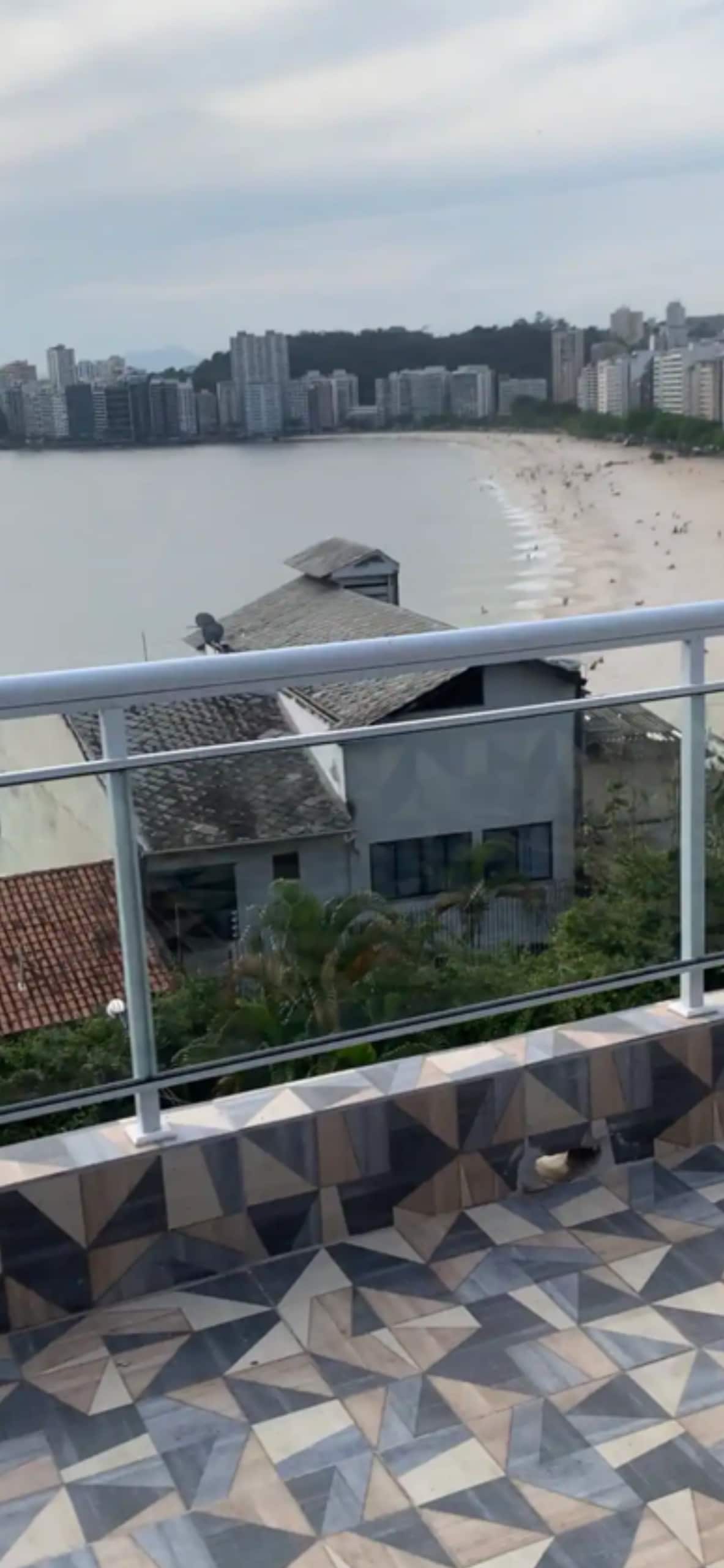 A terrace view overlooking the coastline, showcasing a mix of residential buildings and sandy beach. Gentle waves lap at the shore, framed by greenery below. The railing provides safety while allowing unobstructed views of the scenic landscape, with sunbathers visible in the distance.