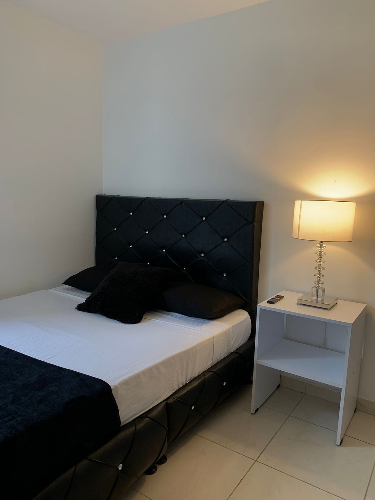 A compact sleeping area is presented, featuring a bed with a tufted black headboard. Two black pillows rest atop a crisp white bedspread. A nightstand stands beside the bed, topped with a lamp that emits a warm glow, complementing the light-colored walls.