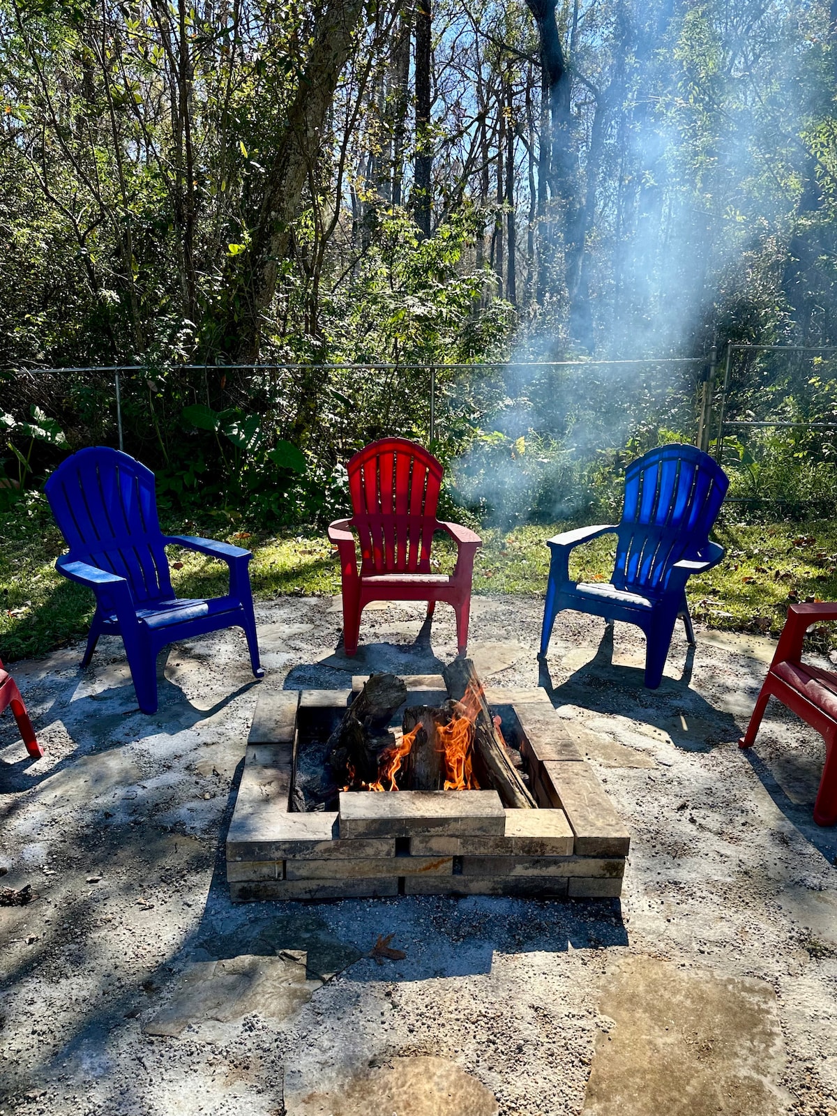 A circular fire pit surrounded by four brightly colored adirondack chairs in shades of blue and red. Smoke gently rises from the crackling flames, with a natural setting of trees and greenery in the background, enhancing the outdoor ambiance.