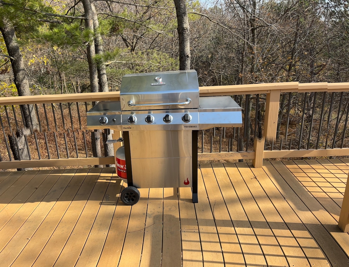 A stainless steel grill is positioned on a wooden deck, surrounded by tall trees. The deck features wooden flooring and a railing that provides a natural setting for outdoor cooking and dining.